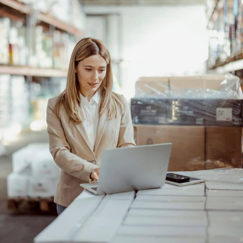 Businesswoman using a laptop in a warehouse to manage production and inventory through an MRP system.